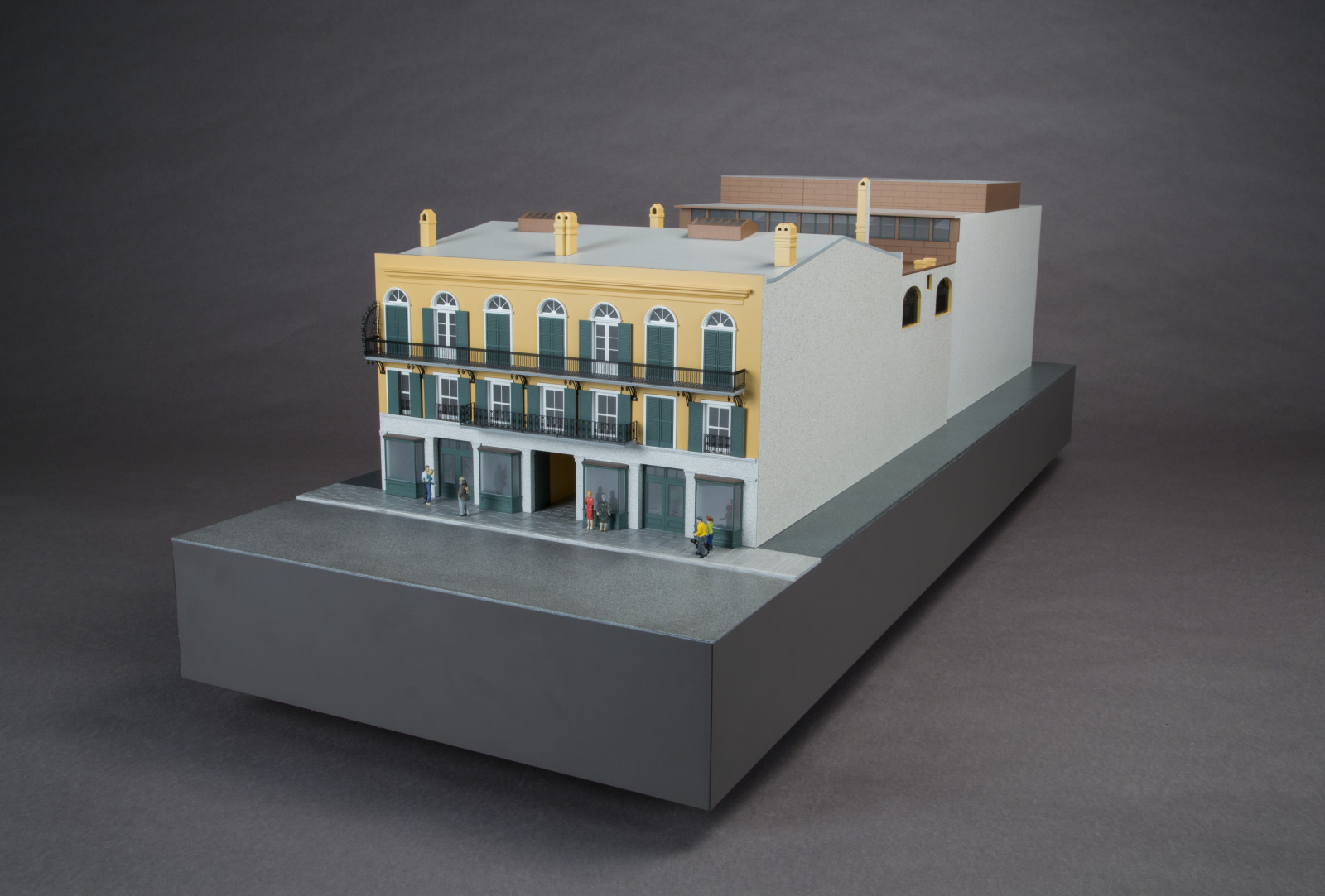 Scale model of a yellow and white two-story building with balconies and arched windows. The structure sits on a gray platform with small figures at street level, against a dark background.