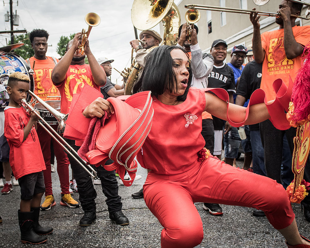 A woman in red dances energetically on a street, holding a hat. Behind her, a brass band plays instruments, with a mix of adults and a child. The scene captures a lively outdoor celebration with spectators in the background.