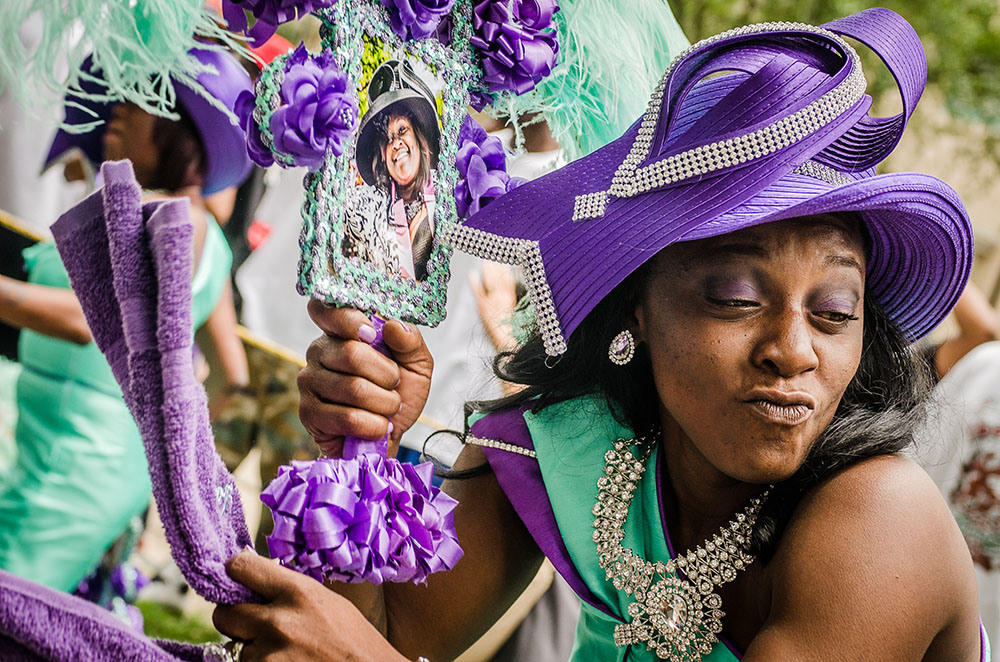 A woman in a vibrant purple and turquoise outfit, complete with an elaborate hat, makes a playful expression. She holds a decorated frame with a photo inside, surrounded by purple fabric and flowers.
