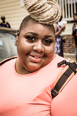 A person smiling, wearing a bright coral shirt and a backpack strap over one shoulder. They have a unique hairstyle with blonde braids styled in a high bun. The background shows blurred figures and part of a house.