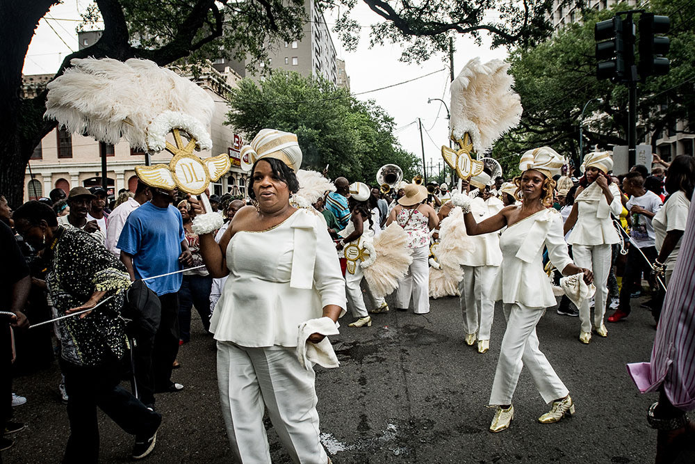 A lively street parade features people in white outfits and feathered headpieces, dancing and holding decorative parasols. The group moves energetically, with a crowd watching from the sides. Trees and buildings are visible in the background.