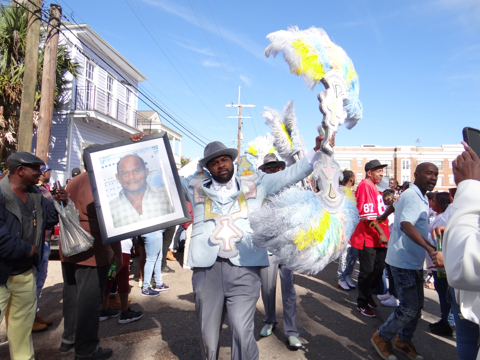 In a vibrant New Orleans street parade, a man dressed in a tailored blue suit with intricate embellishments proudly carries a framed portrait of a loved one. He waves a large, decorated feathered accessory high, surrounded by a festive crowd, as the procession moves through the neighborhood.