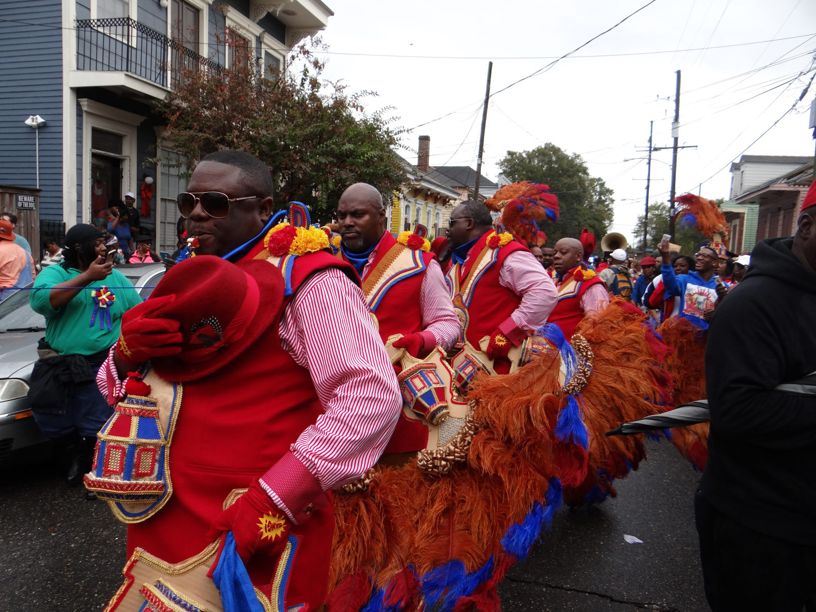 In a lively second-line parade, men dressed in red, gold, and blue strut through the streets, their outfits adorned with embroidered details and flowing feathered fans. The frontman clutches his matching red hat and blows a whistle, while behind him, the procession continues with musicians and dancers.