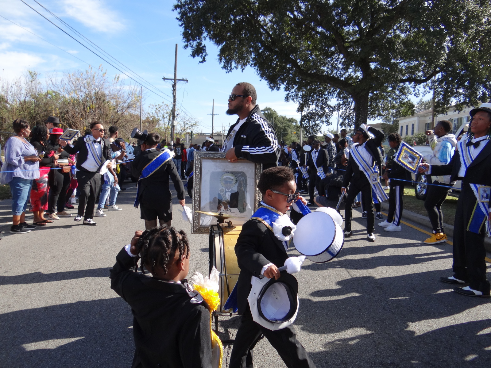 A child in a formal black suit with blue trim walks in a parade, gripping a drum and adjusting his sunglasses. Behind him, a group of performers in matching attire carry framed portraits, while a man in a black tracksuit pushes a cart holding a framed image, likely a tribute. The crowd watches as the procession moves down the sunlit street.