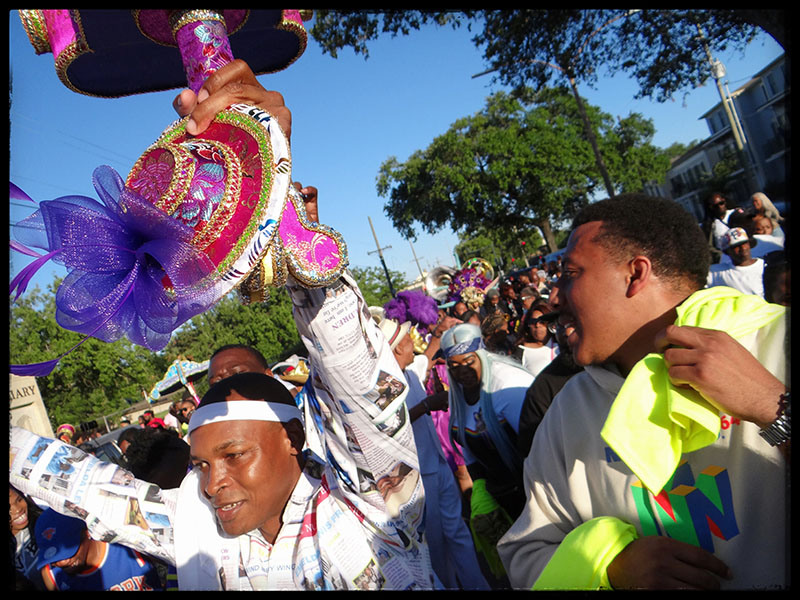 A lively street parade with joyful participants. A man in a printed outfit and headband holds a colorful parasol, while another in a white shirt cheers. The crowd behind them joins in the celebration, with trees and buildings in the background.