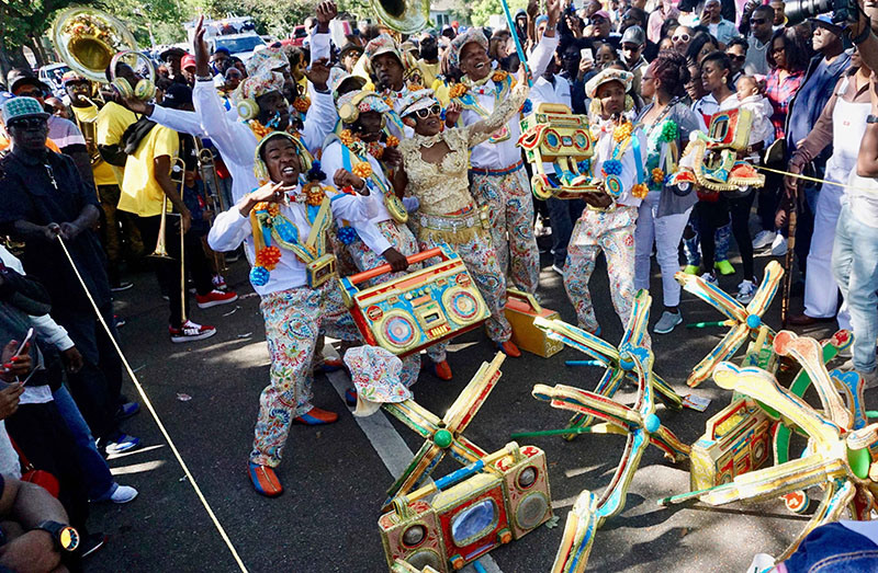 A lively street parade features performers in colorful outfits dancing with large, decorated boombox props. A crowd of spectators surrounds them, capturing the vibrant scene.