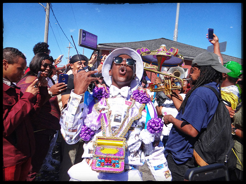 A lively parade scene with a central figure dressed in colorful, decorated attire and holding a hand gesture. Surrounding people take photos and play instruments. The sky is clear, and the atmosphere is festive.