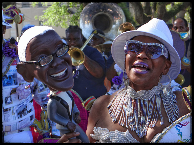 A festive scene with a woman in a white hat and sunglasses smiling, surrounded by musicians playing brass instruments. She is holding a large puppet of a man in glasses and a suit. The background is lively with vibrant colors and joyful expressions.