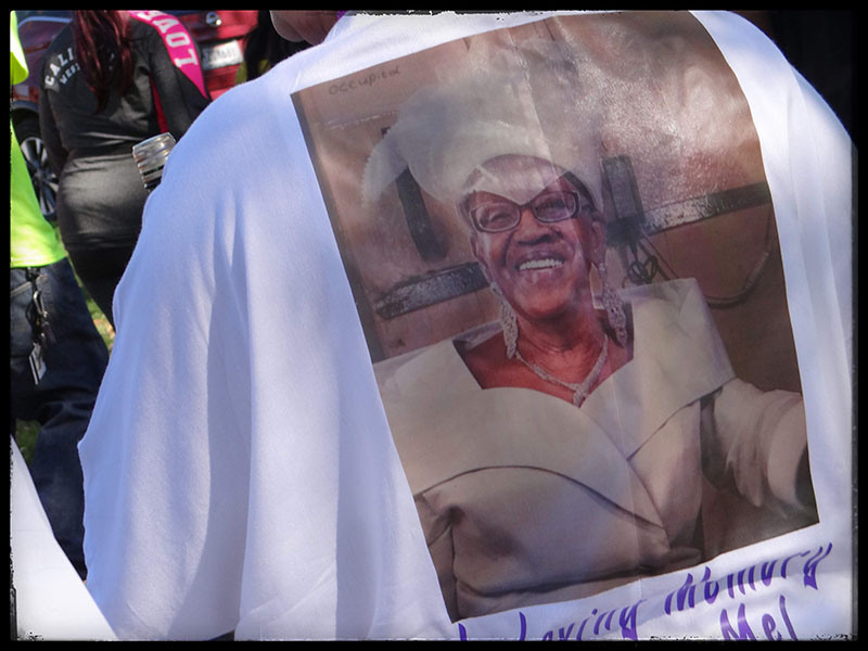 A person wears a shirt with a printed tribute featuring an older woman in a white outfit and hat, smiling. The shirt appears to be part of a memorial gathering, as In Loving Memory text is partially visible at the bottom.