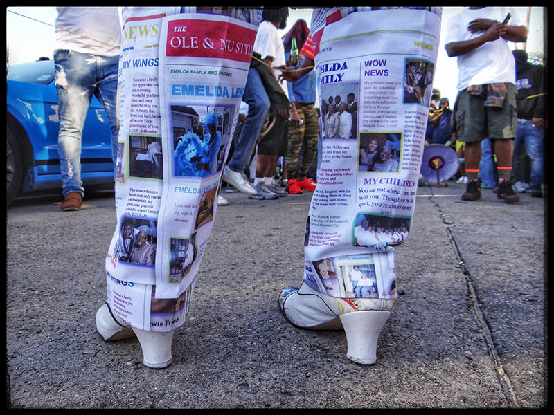 A person wears boots covered in newspaper print, standing on a street. The ground is concrete, and people with casual attire are gathered in the background. A blue car is partially visible on the left side of the image.