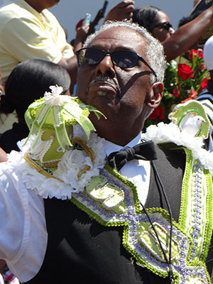 An older man wearing sunglasses and a decorative outfit with green, white, and silver embellishments participates in an outdoor event. He wears a bow tie and white shirt, surrounded by people.