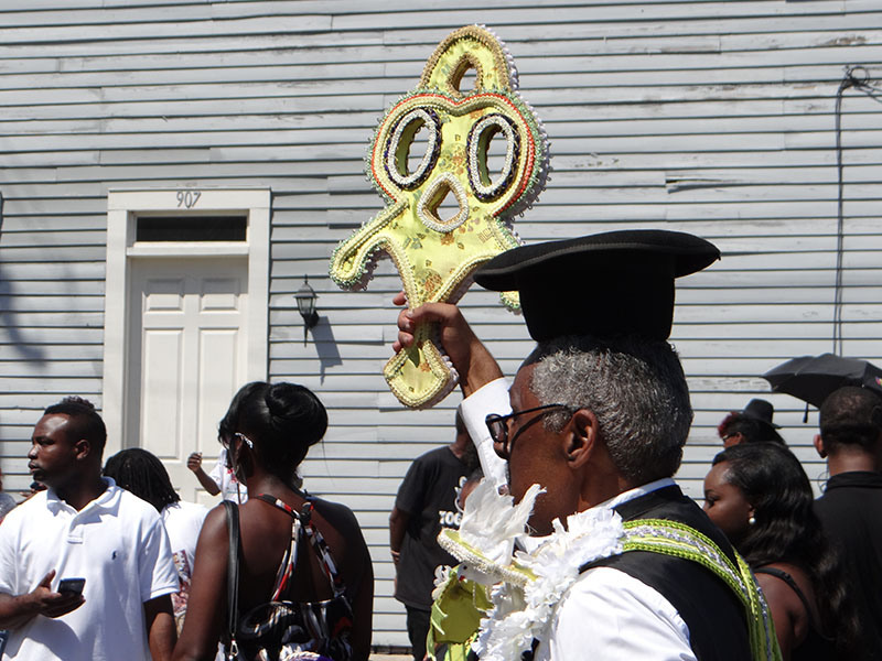 A man in a graduation cap holds a large, decorated yellow key in a crowd during a sunny outdoor event. People around him are dressed in casual attire, and a wooden building is in the background.