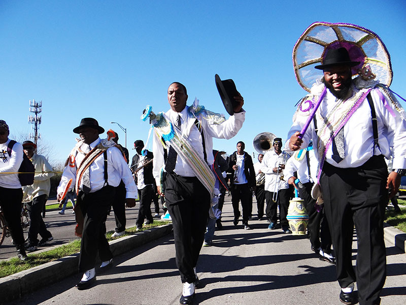 A group of men in colorful costumes and hats parade energetically down a street. One man, in the center, holds his hat in hand. The sky is clear and blue, and spectators watch from the side.
