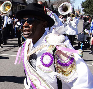 A person in a festive outfit with colorful beads and white feathers dances energetically during a street parade. A brass band with trumpets and tubas follows in the background under a clear blue sky.