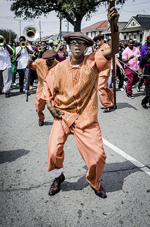 A man in an orange outfit and hat dances energetically on a street during a parade. A brass band and other participants are seen in the background, with houses lining the street.
