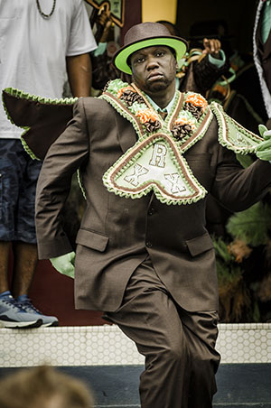 A man dressed in an elaborate brown suit with green and white accents, including gloves, a hat, and a decorated cape. He appears to be dancing or performing on a staircase, surrounded by onlookers.