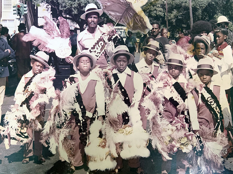 A group of children and an adult dressed in matching costumes with feathers and hats stand together on a street during a parade. The adult holds a decorative umbrella. Trees and other people are visible in the background.
