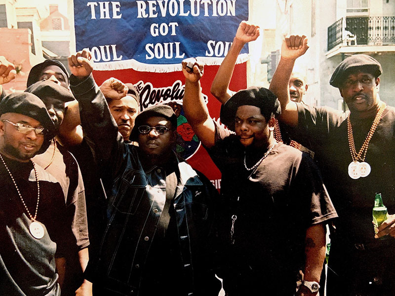 A group of people stand together with raised fists, wearing black attire and berets. A banner behind them reads The Revolution Got Soul. They pose outdoors, demonstrating solidarity and unity.