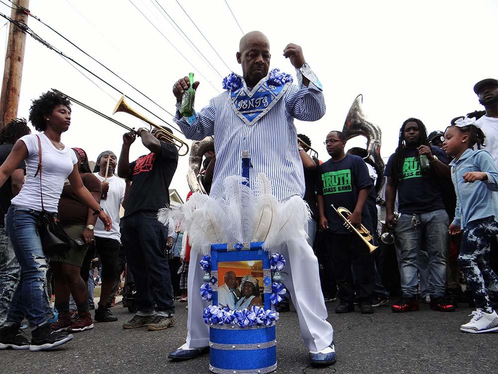 Man dressed in blue and white dances joyfully at a street celebration, surrounded by musicians playing trumpets and trombones. Others look on, wearing casual clothes. A decorated drum with a photo and feathers stands in front of him.