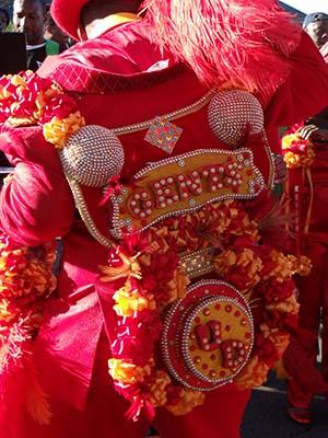 Person wearing an elaborate red costume adorned with sequins, beads, and colorful floral decorations. The back features intricate patterns and the word QUEENS in bold letters.