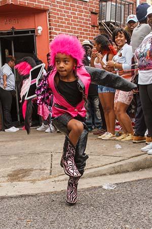 A young girl energetically dances in the street wearing a vibrant pink outfit with matching furry hat and zebra-patterned boots, while onlookers watch and smile in front of a brick building.