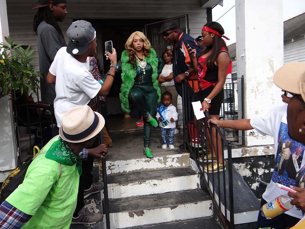 A person in a green outfit and blonde wig walks down steps, surrounded by people taking photos. A small child stands nearby. The scene is lively, with various individuals in casual attire gathered around, capturing the moment.