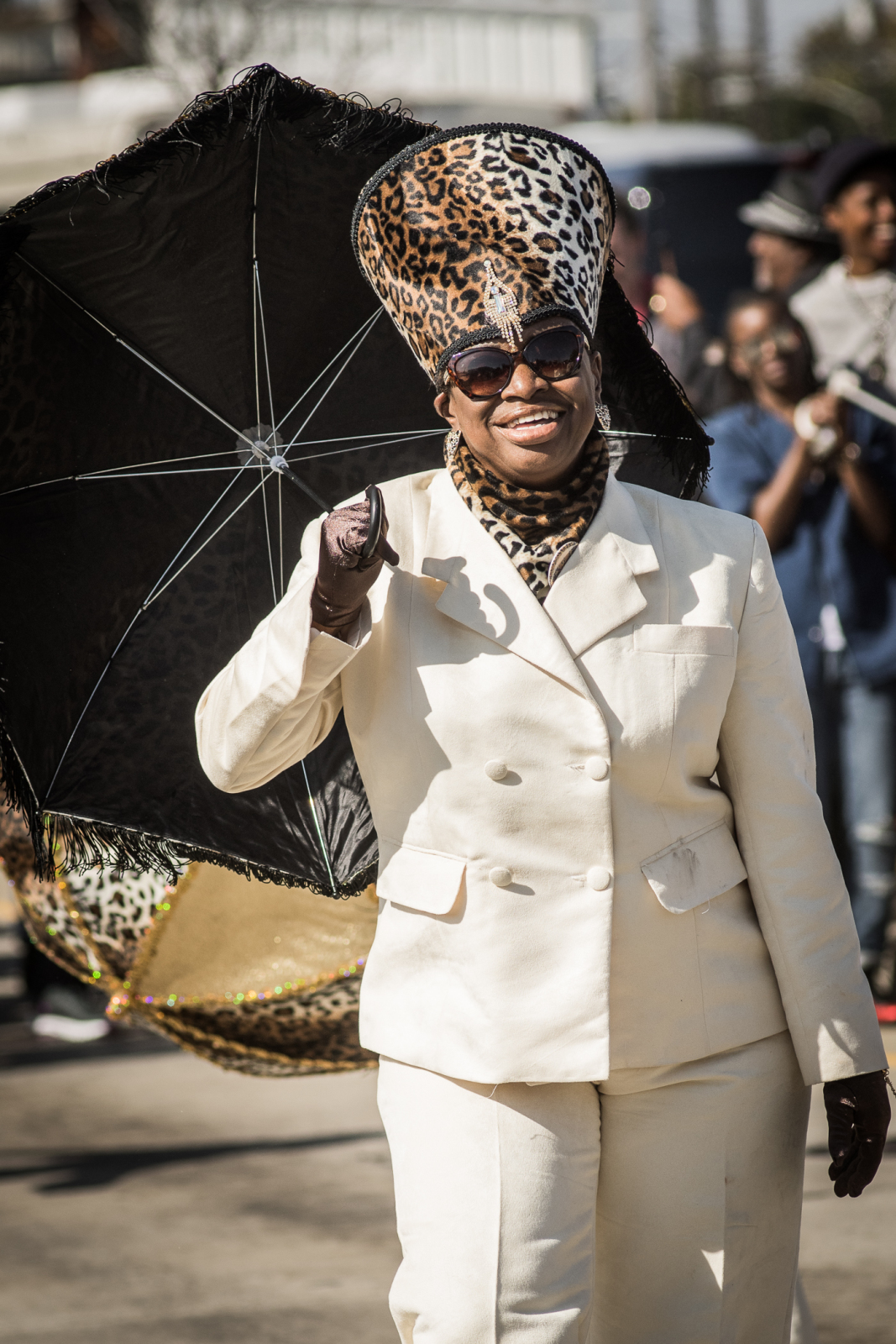 A parade participant in a white double-breasted suit wears a towering leopard-print hat and matching scarf, holding a decorated umbrella with a joyful expression.