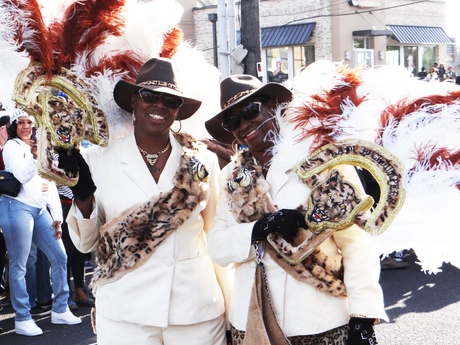Two women in matching white suits and leopard-print sashes smile while holding ornate feathered parade props with tiger faces. A crowd watches in the background.