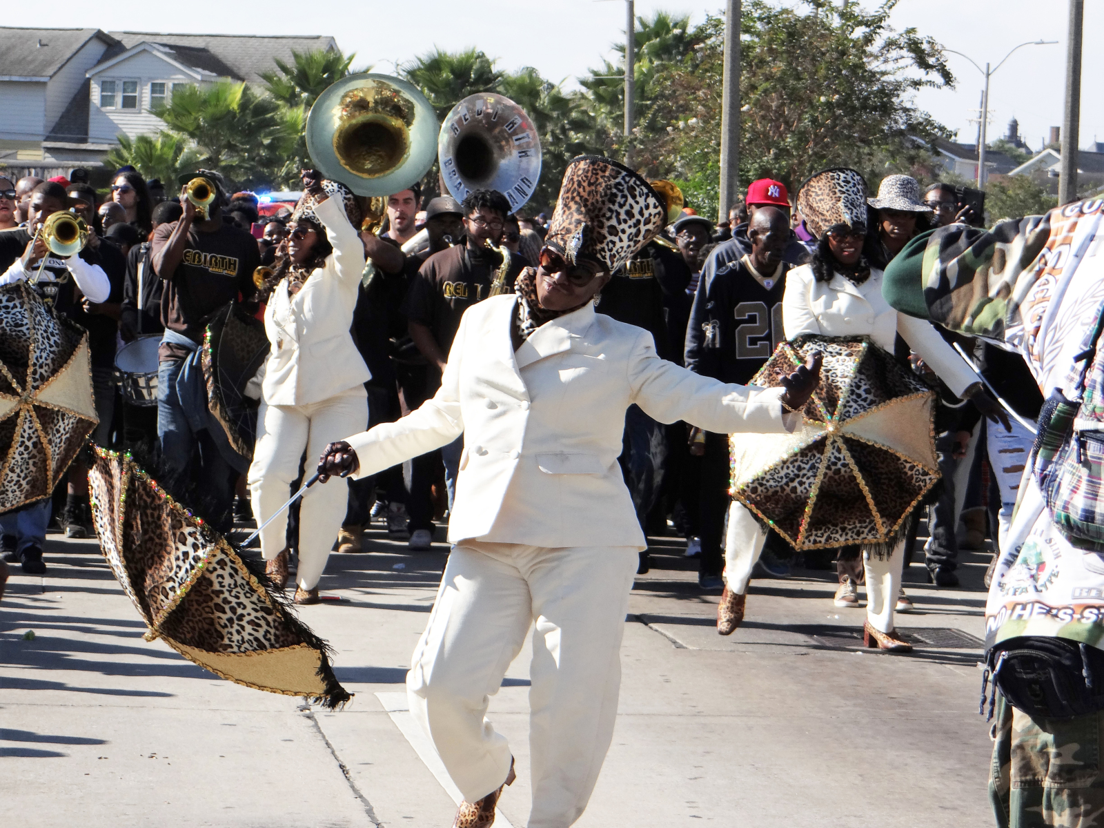 A second line parade moves through the street, led by a woman in a white suit and leopard-print accessories dancing with a coordinating umbrella, while a brass band plays behind her.
