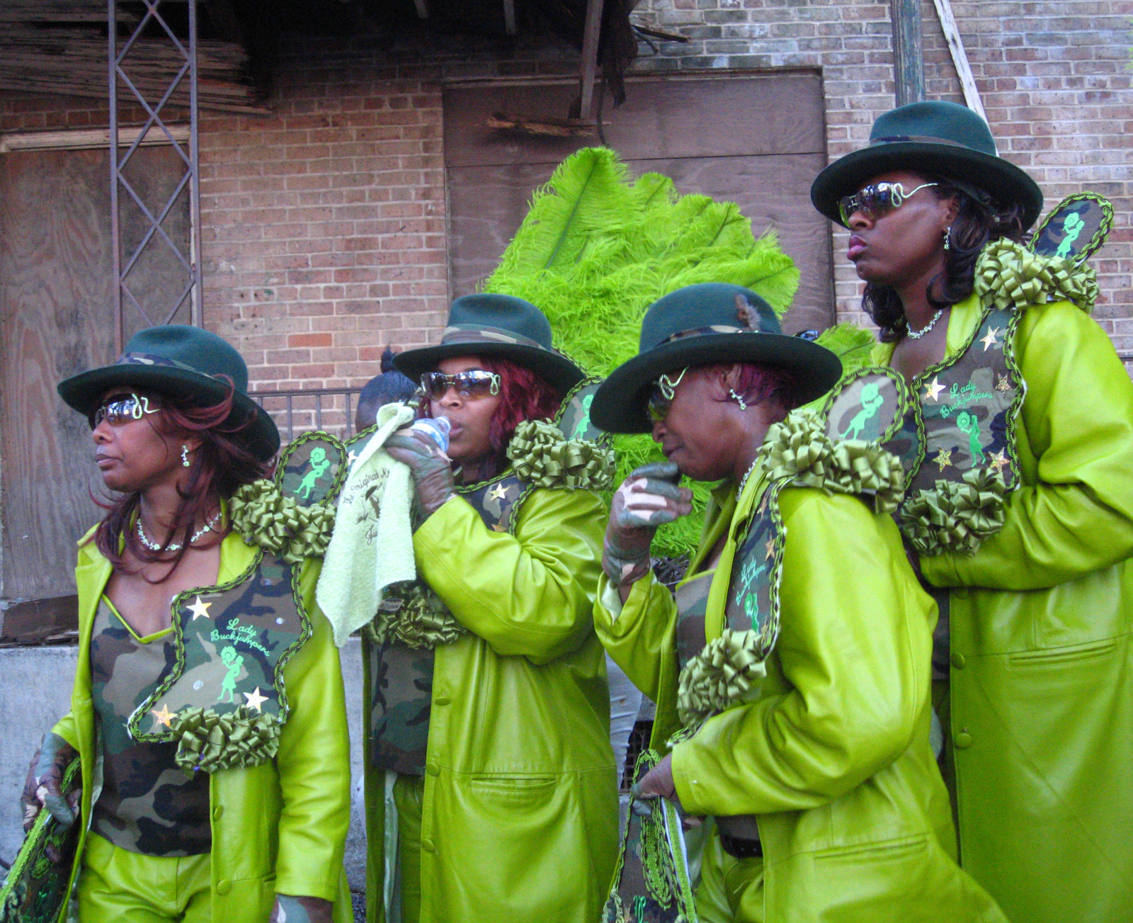 A group of women dressed in matching neon green and camouflage outfits stand together during a parade, wearing green hats, sunglasses, and decorative shoulder adornments. One woman drinks water while another wipes her face with a towel.