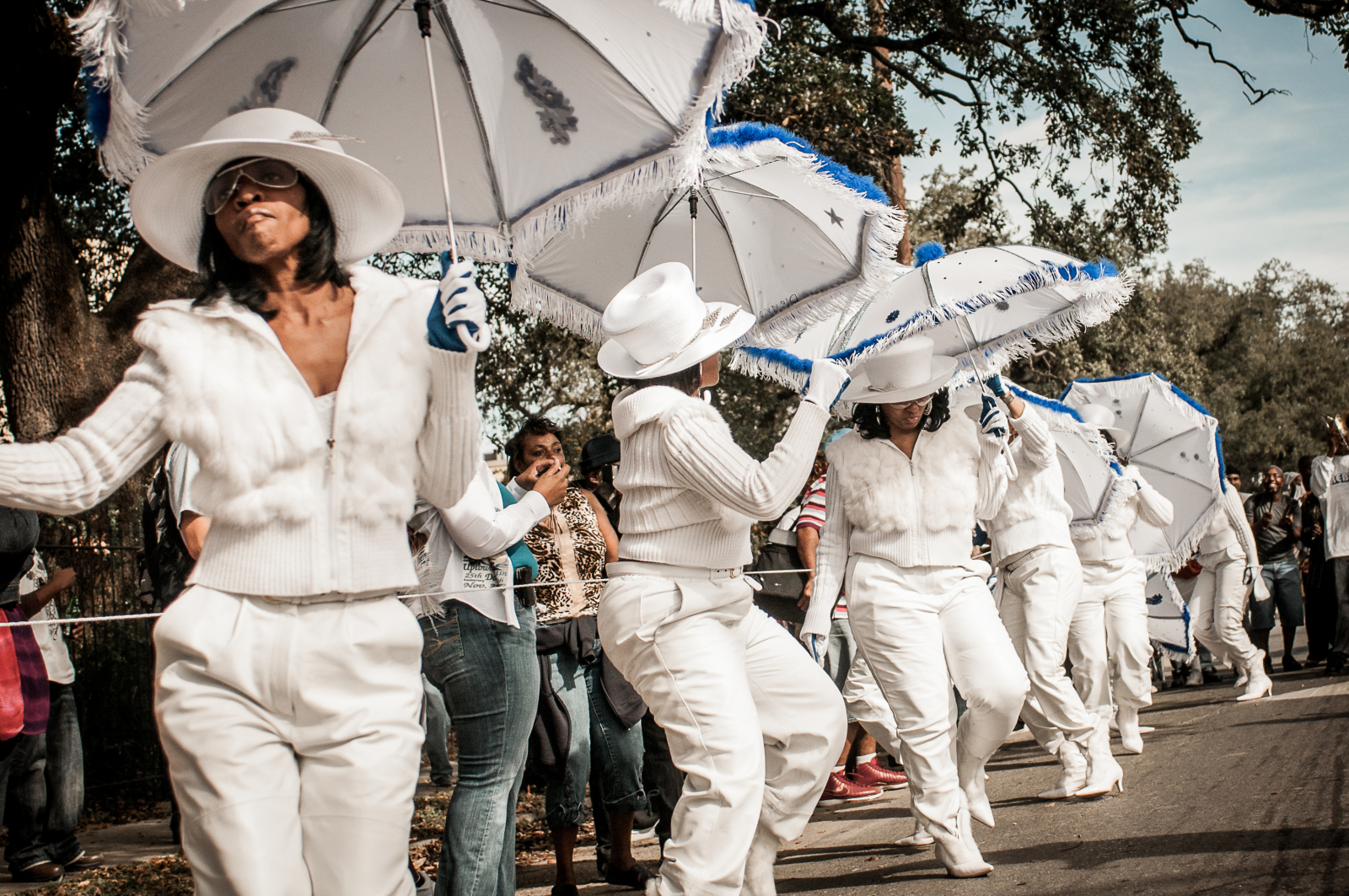 Parade performers dressed in elegant all-white outfits, including wide-brimmed hats, fur-trimmed jackets, and gloves, dance in unison during a parade. They hold decorated white umbrellas with blue fringe as they step to the rhythm, surrounded by onlookers.