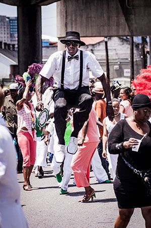 A vibrant street parade with a man on stilts wearing a hat, bow tie, and suspenders. Colorful participants dance and march, holding festive decorations. Urban buildings and poles are visible in the background.