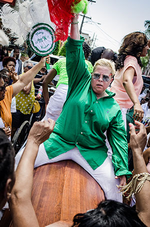 Person in a green shirt and white pants riding a wooden object, surrounded by a lively crowd. Others are holding decorative items, and the scene appears festive, possibly part of a cultural celebration or parade.