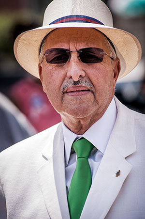 An elderly man wearing a white suit, a green tie, and a white hat with a blue and red band. He has sunglasses and a small pin on his lapel. The background is softly blurred.