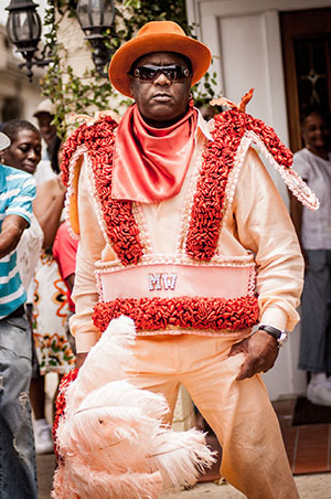 A man in an elaborate orange costume with red and white fringes stands confidently. He wears sunglasses and a hat, holding a decorated accessory. People and a building are in the background.