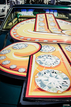 A colorful sign with large, decorative faux diamonds and orange hues is placed on the hood of a green car.