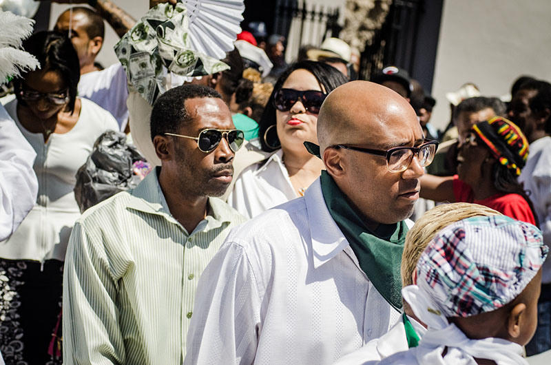 A group of people wearing white clothing participate in an outdoor event. Some hold fans, and one person wears sunglasses. The atmosphere appears lively and celebratory, with a diverse crowd in the background.