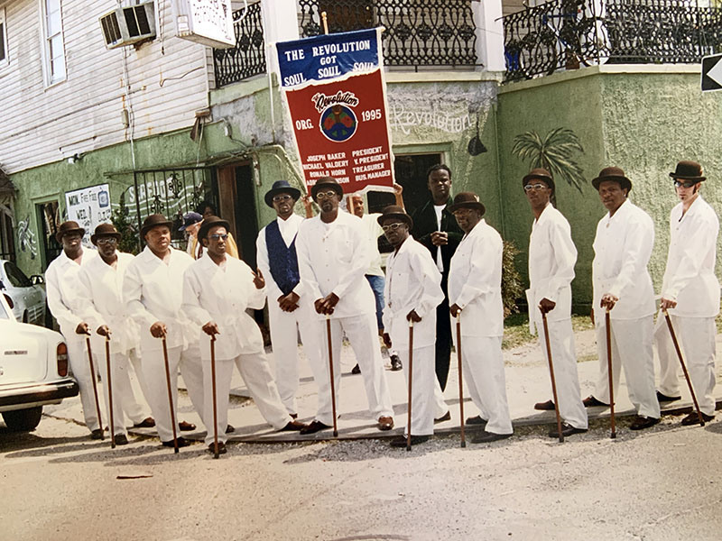 A group of men in white suits and hats stand together on a street. They hold walking sticks and are posed in front of a sign that reads The Revolution Got Soul. 1995. A vintage car is partially visible on the left.
