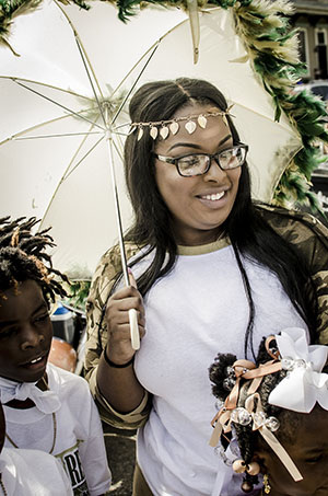 A woman in glasses, holding a decorative umbrella, stands outdoors with two children. She wears a headband with shell-like decorations and a white shirt. The children have braided hair with bows. The scene is cheerful and festive.