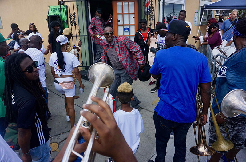 A vibrant street celebration with people dancing and playing brass instruments. One man in a colorful outfit stands in the center, surrounded by others. Musicians are holding trumpets and trombones, creating an energetic atmosphere.