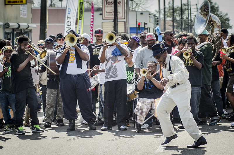 A lively street performance featuring a brass band with trumpets and trombones. A man in a white suit and hat energetically dances in front of the musicians, while a crowd gathers to watch. The setting is urban with storefronts in the background.