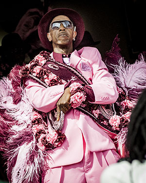 A person wearing a bright pink suit and a matching hat stands confidently with arms crossed. They are adorned with colorful feathers and decorations, and wear sunglasses, creating a striking and stylish appearance.