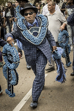 An elderly man wearing a blue patterned suit and sunglasses is leading a lively parade. He holds ornate, blue decorations and is surrounded by other parade participants, some holding musical instruments.