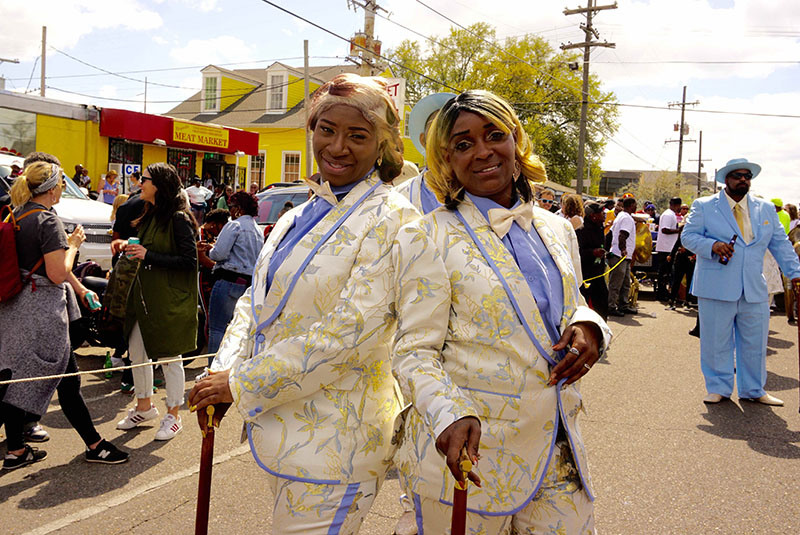 Two people stand back-to-back, both wearing elaborate matching suits with floral patterns and holding canes. Theyre part of a lively street parade, surrounded by a crowd enjoying the event. One person is in a light blue suit in the background.