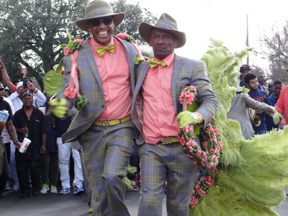 Two men in matching plaid suits, pink shirts, and green bow ties dance energetically in a street parade. They are adorned with floral garlands and surrounded by a lively crowd and trees in the background.
