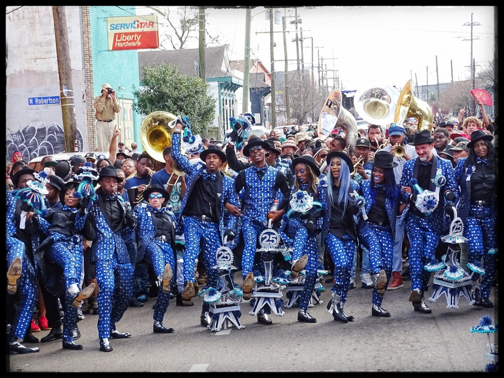 A vibrant parade features a group of performers dressed in matching blue and black polka dot outfits, holding decorated umbrellas. A lively crowd watches as they dance energetically down the street, with buildings and instruments visible in the background.
