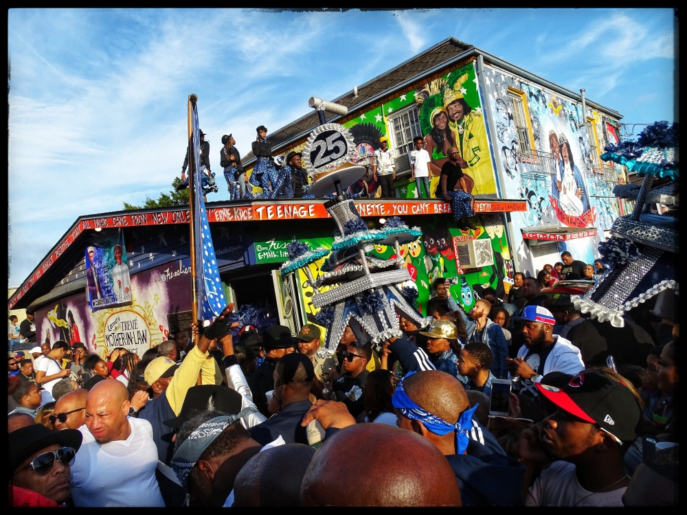 A vibrant street scene shows a large crowd gathered in front of a colorful, graffiti-covered building. People are wearing casual clothes and hats, celebrating with enthusiasm. The sky is clear, suggesting a sunny day.
