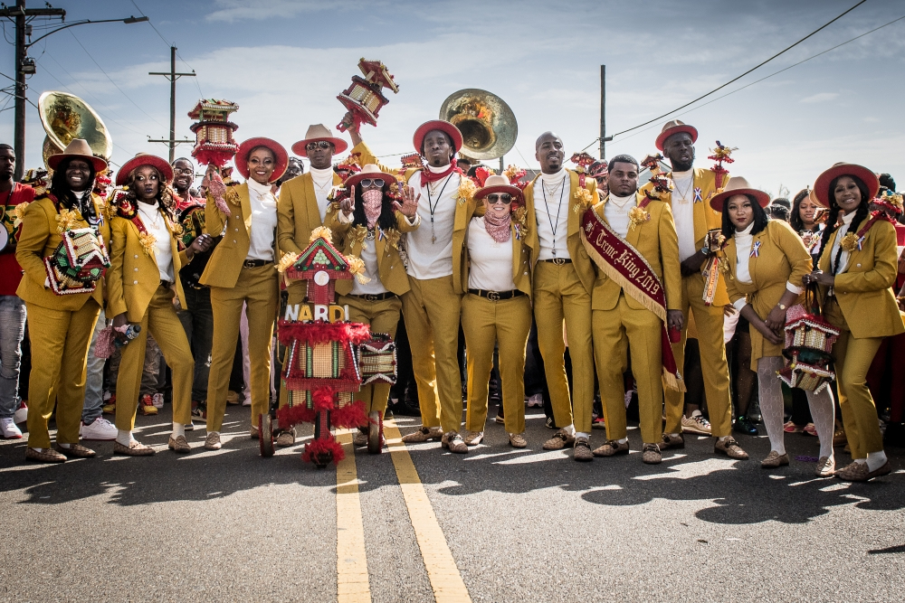 A group of people in matching yellow suits stand on a street holding instruments and colorful decorations. They are participating in a festive parade under a clear sky.