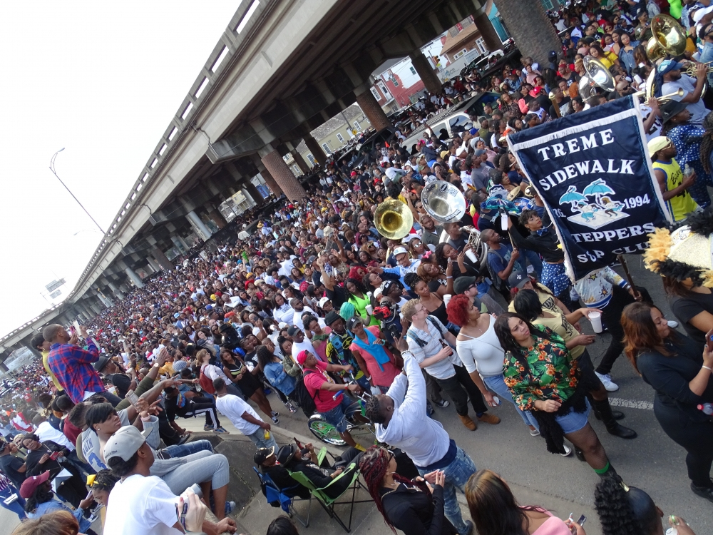 A large crowd gathers under a highway overpass for a festive parade. People are celebrating with music and dancing. A banner reads Treme Sidewalk Steppers SOP. The atmosphere is lively and colorful.