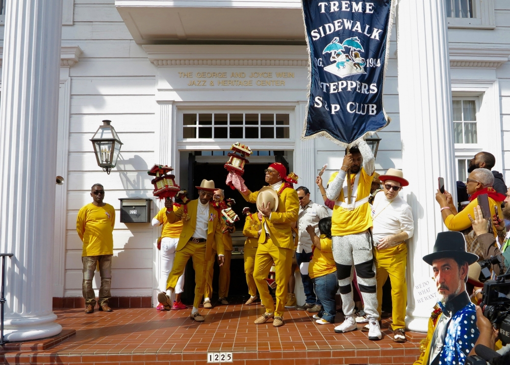 A group of people dressed in vibrant yellow outfits and red accessories stand on the steps of a building with white columns. They hold small decorated floats. A banner reads Treme Sidewalk Steppers S&P Club. A crowd gathers to watch.